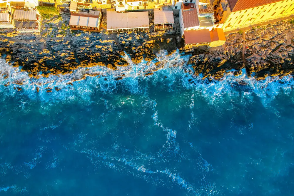 onde che si infrangono su costa rocciosa con edifici.