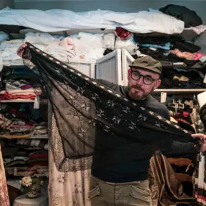 Uomo mostra scialle nero ricamato, in stanza piena di tessuti.