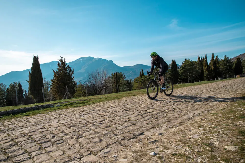 Ciclista in bici da corsa su strada acciottolata, sfondo montuoso.