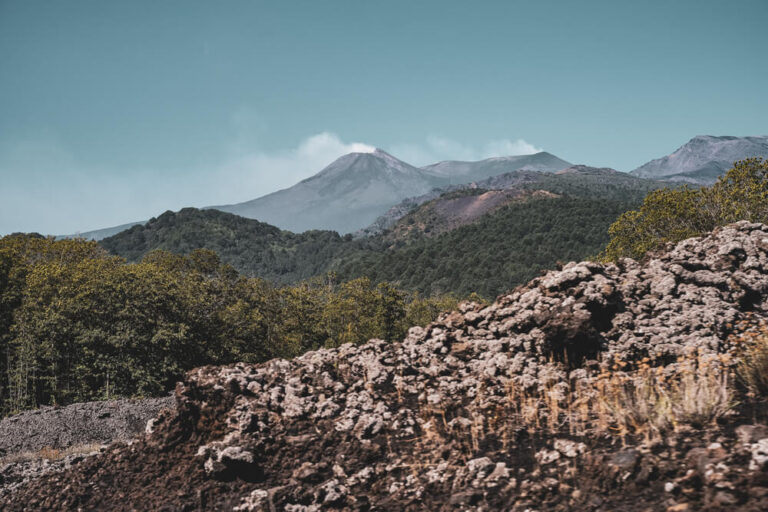 Vulcano in lontananza, vegetazione lussureggiante e rocce vulcaniche in primo piano.